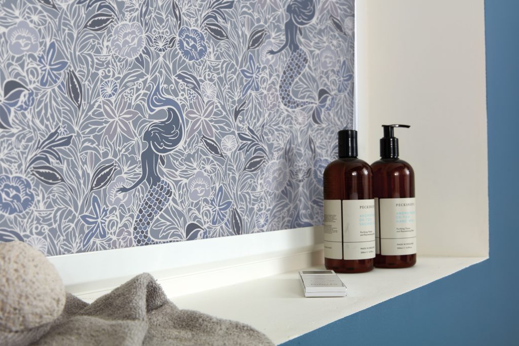 Close‑up of a bathroom window with an indigo roller blind featuring a detailed mermaid and floral pattern, styled with amber toiletry bottles and soft towels on the white sill.